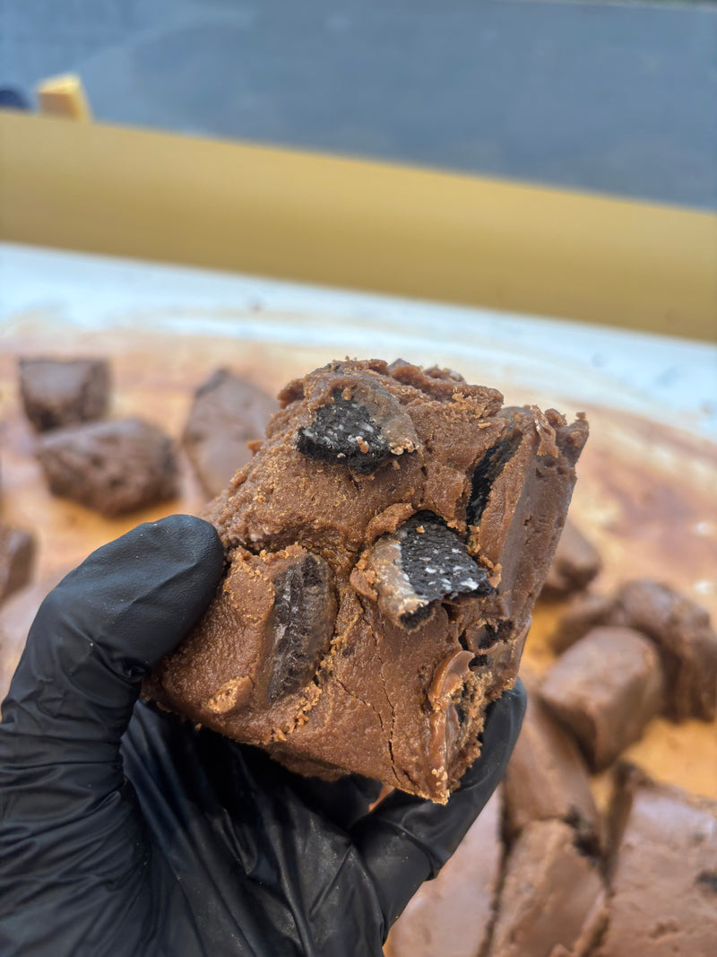 Chocolate fudge with chocolate chips held by a gloved hand, with more fudge in the background.